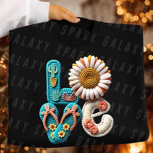 May include: Black sweatshirt featuring the word "LOVE" in a vibrant, crocheted design. The "L" is a blue cactus, the "O" is a daisy, the "V" is flip-flops, and the "E" is decorated with flowers. The background has a repeating pattern of "SPARK GALAXY".