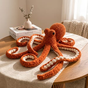 May include: A large, handmade crochet octopus plush toy featuring a vibrant burnt orange exterior and a contrasting cream-coloured underside on its eight curled tentacles. The stuffed creature has two dark, round eyes and a textured, soft yarn finish.