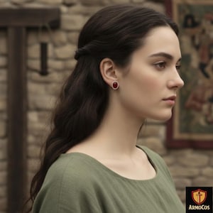 May include: A woman wearing a green dress and red oval earrings. The earrings have a silver setting and a deep red stone. The woman has long, dark hair and is looking to the side. The background includes a stone wall and a framed picture.