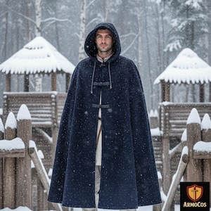 May include: A navy blue hooded cloak with toggle closures. The cloak is worn over a white tunic. The background shows a snowy forest and wooden structures. The cloak is from ArmoCos.