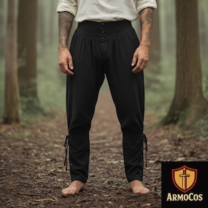 May include: Black medieval-style pants with a high waist and button closure. The pants have a loose fit and are tied at the ankles with laces. The pants are made of a soft, flowing fabric. The Armocos logo is in the bottom right corner.