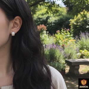 Peut inclure: Gros plan sur une oreille portant une petite boucle d'oreille carrée et texturée. La boucle d'oreille est de couleur claire et neutre. L'arrière-plan est une scène extérieure floue avec de la verdure et un banc en pierre.