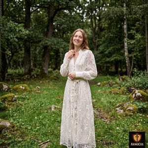 Peut inclure: Longue robe blanche en dentelle avec manches longues et boutonnage sur le devant. La robe est nouée à la taille et portée en extérieur dans un cadre forestier. La robe est de style vintage ou bohème.