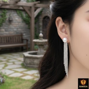 Peut inclure: Boucles d'oreilles composées d'une perle blanche et d'une longue chaîne argentée. Le design classique convient aux tenues formelles ou décontractées. L'image est prise sur un fond extérieur flou.
