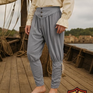 Op de afbeelding: Lichtgrijze piratenbroek met knoopdetails en veter manchetten. De broek wordt gecombineerd met een crèmekleurig shirt met lange mouwen. De outfit is geschikt voor cosplay of historische re-enactments.