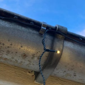 May include: A string of small, warm white LED lights attached to a weathered metal gutter with a silver plastic clip. The dark wire runs along the length of the gutter, with one illuminated bulb hanging down against the textured, gray surface.