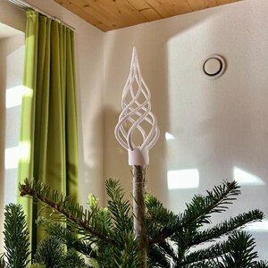 Op de afbeelding: Een witte, spiraalvormige, vlamvormige piek voor in de boom met een open, kooiachtige structuur van gebogen, elkaar kruisende lijnen. Het ornament is bevestigd op de centrale, verticale tak van een groene naaldboom.