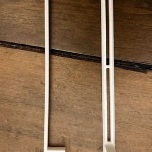 May include: Two white plastic components, each featuring a long, narrow vertical bar. The left piece has a triangular support bracket at the base, while the right piece includes a central vertical slot and a small, protruding hook-like attachment.