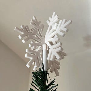Könnte beinhalten: Ein weißer Schneeflocken-Weihnachtsbaumschmuck. Der Schmuck besteht aus weißen, geometrischen Formen, die wie eine Schneeflocke angeordnet sind. Der Schmuck ist auf einer weißen Stange montiert und befindet sich auf einem grünen Weihnachtsbaum.