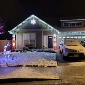 Op de afbeelding: Een huis versierd met witte ijspegelverlichting langs de dakrand, een verlicht sneeuwvlokornament en rood-witte zuilverlichting. Een boom met veelkleurige lichtjes en een wit rendier van draad staan op het gazon voor de woning.