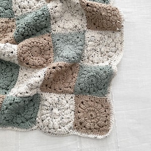 May include: A handmade crochet blanket composed of square motifs in a repeating pattern. The squares feature a circular floral design in shades of sage green, tan, and cream with black flecks throughout the yarn, finished with a scalloped edge.