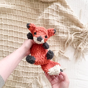 May include: A handmade crochet fox toy in shades of orange, gray, and white. The fox has a detailed design with a fluffy tail and small ears. The toy is held against a textured, cream-colored blanket.
