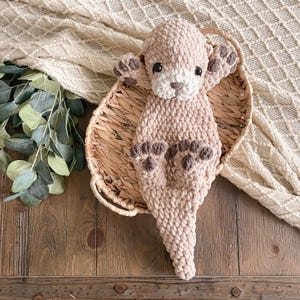 May include: A plush, crocheted otter toy made from soft, light beige yarn. The otter features a white muzzle, dark brown embroidered nose, dark plastic eyes, and dark brown paw pads on its four limbs. The body has a textured, fuzzy appearance.