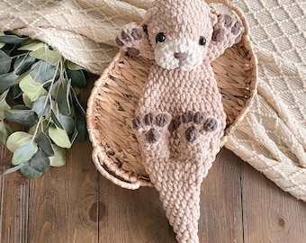 May include: A plush, crocheted otter toy made from soft, light beige yarn. The otter features a white muzzle, dark brown embroidered nose, dark plastic eyes, and dark brown paw pads on its four limbs. The body has a textured, fuzzy appearance.