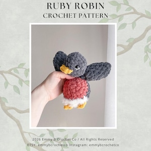 Op de afbeelding: Een gehaakt vogel speelgoed met de tekst "RUBY ROBIN CROCHET PATTERN". De vogel heeft grijze vleugels en kop, een rode borst, witte buik en gele snavel en poten. De vogel wordt in een hand gehouden.