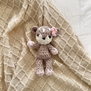 May include: A small, handmade crochet deer plush toy featuring a light brown body, a cream-colored face, and a small pink flower accent on its head. The deer has dark eyes, a dark nose, and light pink accents on its ears, hands, and feet.