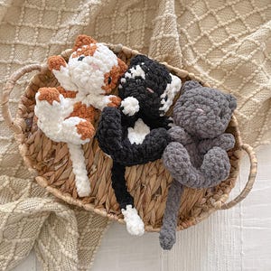 May include: Three plush cat toys made of soft, chunky yarn resting in an oval woven basket. One cat is white with orange patches, one is black with white markings, and the third is solid gray. Each cat has small ears, a long tail, and stitched eyes.