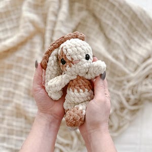 May include: A small, handmade crochet plush toy shaped like a rabbit with long, floppy ears. The toy is crafted from soft, chunky yarn in alternating patches of cream and light brown, featuring two small black plastic eyes and a stitched nose.