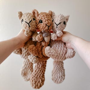 May include: Three plush stuffed animals made of soft, chunky yarn in shades of beige, light brown, and tan. Each animal features embroidered black facial details, small ears, and long, textured tails. The center animal holds a small brown acorn.