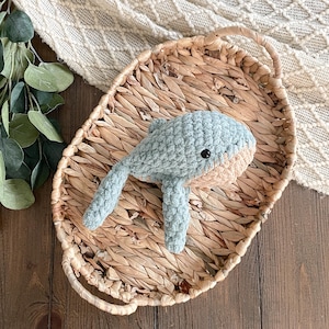 May include: A crocheted whale toy in light blue and tan, with a black eye, rests in a woven basket. The whale has a textured appearance and is positioned on a diagonal. The basket is oval-shaped and made of natural fibers.