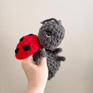 May include: A handmade crochet ladybug plush toy featuring a dark gray textured body and head with a bright red wing section adorned with black spots. The toy has a rounded head with a single visible black plastic eye and small antennae.