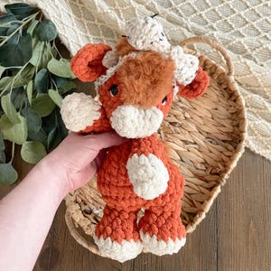 May include: A handmade crochet stuffed animal in the shape of a cow. The plush toy is primarily orange with white accents on the face, feet, and ears. The cow has a fluffy brown patch on its head and a small white crocheted hat.
