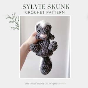 Könnte beinhalten: Ein gehäkeltes Stinktier-Plüschtier in Grau- und Weißtönen, gehalten in einer Hand. Das Stinktier hat einen weißen Streifen auf Kopf und Schwanz, mit einer rosa Nase. Der Text "SYLVIE SKUNK CROCHET PATTERN" steht oben.