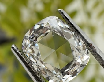 May include: A cushion-cut diamond held by tweezers. The diamond is clear and faceted, reflecting light. The background is a soft green, suggesting a natural setting. The tweezers are silver-colored.