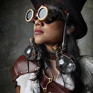 May include: A woman wearing a black top hat, goggles, and a brown leather harness with two glass vials attached. She is wearing a white blouse with lace trim.