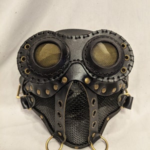 Könnte beinhalten: Eine schwarze Leder-Steampunk-Maske mit goldenen Akzenten und Netz-Augenschutz. Die Maske hat zwei große runde Schutzbrillen mit goldfarbenem Netz und ein kleineres Netz, das Nase und Mund bedeckt.