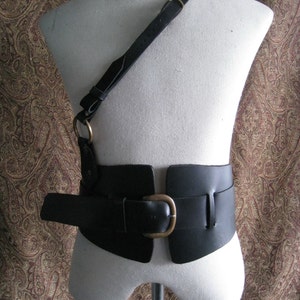 May include: A black leather belt with a large buckle and a matching shoulder strap. The belt is designed to be worn around the waist and the strap can be worn over the shoulder.