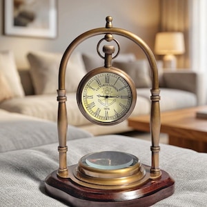 May include: A brass-toned desk clock suspended within an arched frame, featuring a round face with Roman numerals, the text Victoria London, and an anchor graphic with the year 1876. A circular compass is integrated into the dark wooden base.