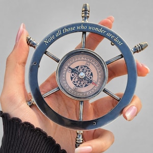 May include: A vintage-style compass in the shape of a ship's wheel. The compass has a dark metallic finish with the inscription "Note all those who wonder are lost" around the rim. The compass face features a detailed design with directional markings.