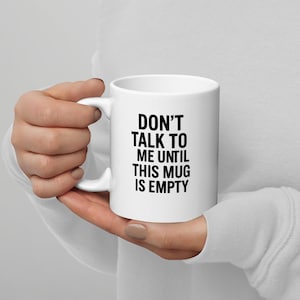 May include: White ceramic mug with the text "DON'T TALK TO ME UNTIL THIS MUG IS EMPTY" in black, held by a person. The mug has a standard handle and a glossy finish. The text is centered on the mug.