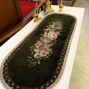 May include: An oval table runner in dark green velvet featuring a central floral tapestry design with pink, cream and green blossoms. The runner is bordered by a dark floral pattern and finished with a scalloped, light beige lace trim edge.