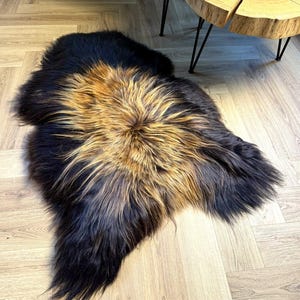 Può includere: Un tappeto in finta pelliccia nera e marrone con un'area centrale color oro-marrone. Il tappeto è posizionato su un pavimento in legno chiaro, con due tavolini in legno con gambe in metallo nero sullo sfondo. I tavoli hanno venature naturali.