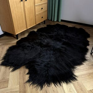 Puede incluir: Una alfombra grande de piel sintética negra con una textura peluda. La alfombra tiene forma irregular y está sobre un suelo de madera marrón claro. Un armario de madera con pomos negros y una cortina verde están en el fondo.