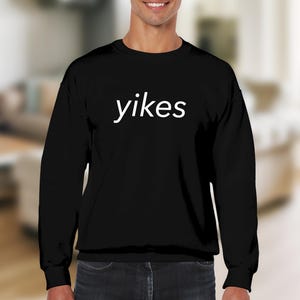 Puede incluir: Sudadera negra de manga larga con la palabra "yikes" en blanco centrada en el pecho. La sudadera tiene cuello redondo y está hecha de un material suave. El fondo está borroso, sugiriendo un entorno interior.