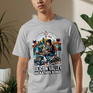 May include: A light gray t-shirt featuring a colorful illustration of a person working on a laptop in front of a cityscape with palm trees and the Golden Gate Bridge. The text "SILICON VALLEY HACKATHON NOMAD" is below the illustration.