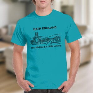 May include: A bright turquoise short-sleeve t-shirt featuring a black line art graphic of the Bath, England skyline. The text "BATH ENGLAND" appears above the illustration, and the phrase "Tea, History & a Little Luxury" is printed below it.