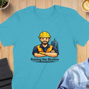 May include: A light blue t-shirt featuring a cartoon illustration of a construction worker wearing a yellow hard hat and safety vest, with arms crossed. The text "Raising the Skyline" is below the image.