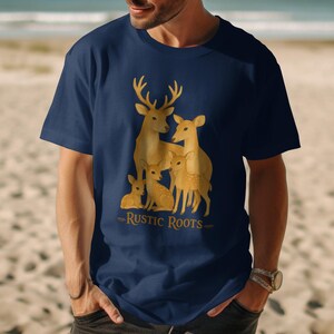 May include: Navy blue t-shirt featuring a golden illustration of a deer family with the text "Rustic Roots" below. The design includes a buck, doe, and fawns. The shirt is made of a soft material.