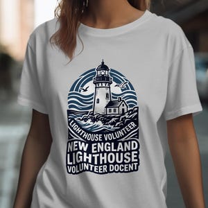 May include: White t-shirt with a navy and white graphic of a lighthouse, waves, and seagulls. Text reads "Lighthouse Volunteer" and "New England Lighthouse Volunteer Docent".