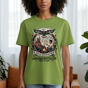 Puede incluir: Camiseta verde claro con un diseño gráfico. El diseño presenta un águila, dos figuras junto a una hoguera y el texto "NATIVE RESERVATION LANGUAGE REVITALIZER". La camiseta tiene mangas cortas y cuello redondo.