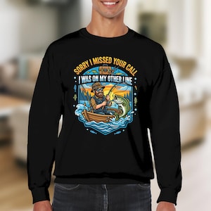 Könnte beinhalten: Schwarzes Sweatshirt mit einem Cartoon-Design eines Fischers in einem Boot beim Angeln. Der Aufdruck enthält den Text "SORRY I MISSED YOUR CALL I WAS ON MY OTHER LINE". Die Grafik ist von Wasser und Fischen umgeben.