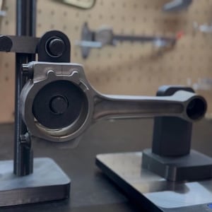 May include: A metallic gray connecting rod, a component of an engine, is displayed on a black stand. The rod is held in place by a black metal fixture. The background shows tools and a pegboard.