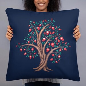 May include: A navy blue cushion with a detailed illustration of a tree. The tree has a brown trunk, green leaves, and red fruit. The tree is centered on the cushion, creating a decorative accent for home decor. The cushion is held by a person.
