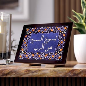 Könnte beinhalten: Dekorative Tafel mit arabischem Text und ornamentalem Blumenrand. Die Tafel hat eine blaue Mitte mit weißer Schrift, umrahmt von einem dunkelbraunen Rand und wird von Holzständern getragen. Das Design umfasst orangefarbene und violette Blumenakzente.