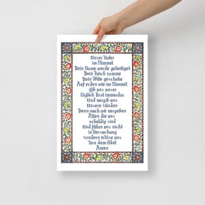 May include: A framed print displaying the German version of the Lord's Prayer. The text is in a decorative font, framed by a floral border with red, yellow, and blue accents. The print is held by a person.