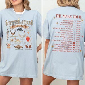 a woman wearing a light blue t-shirt with a graphic design featuring various symbols and text related to the Maas Tour.